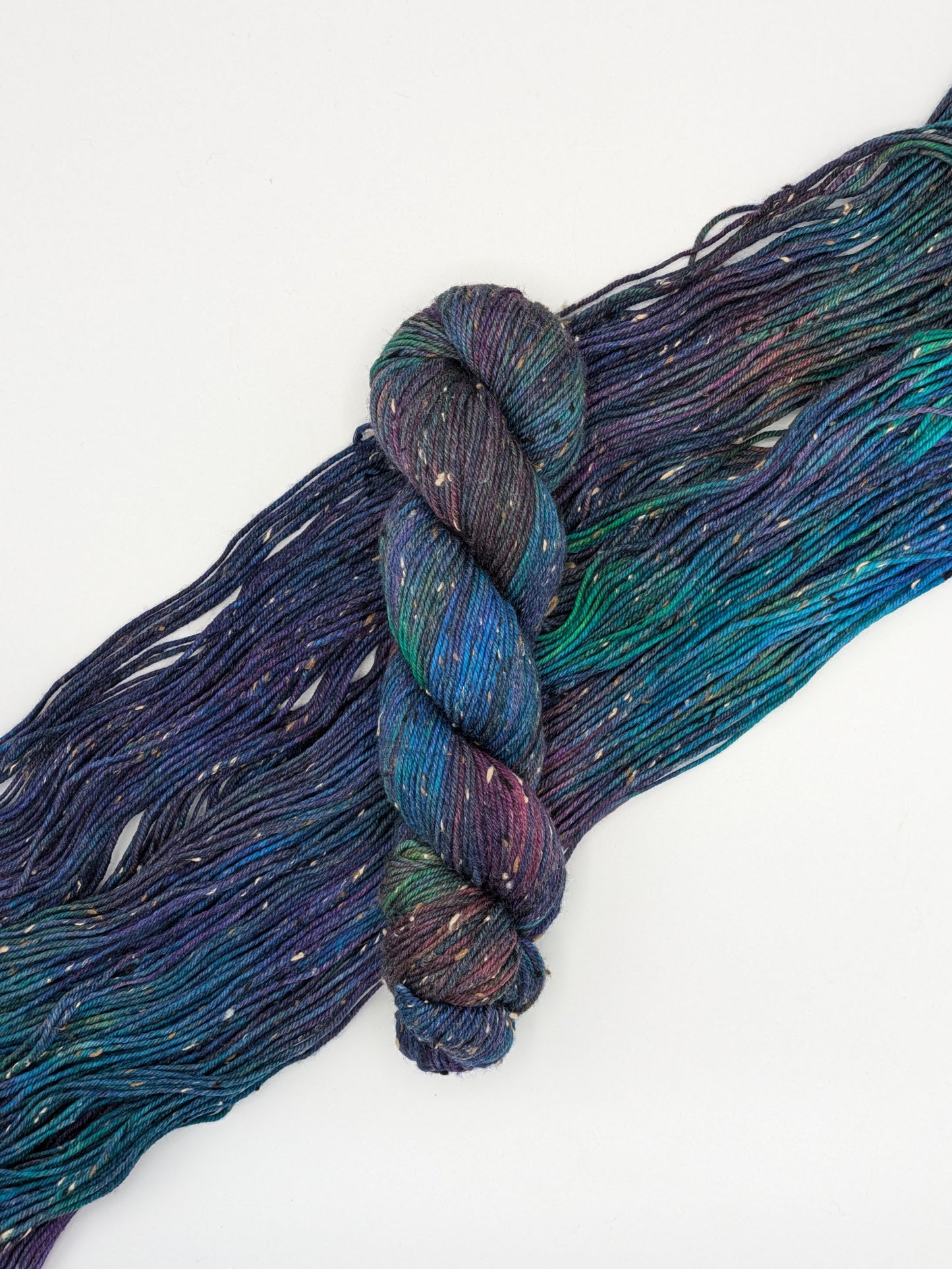 A skein of Galaxy Tweed DK weight yarn laying on a skein spread out to show it's jewel toned colors including blues, green, purples, greens.