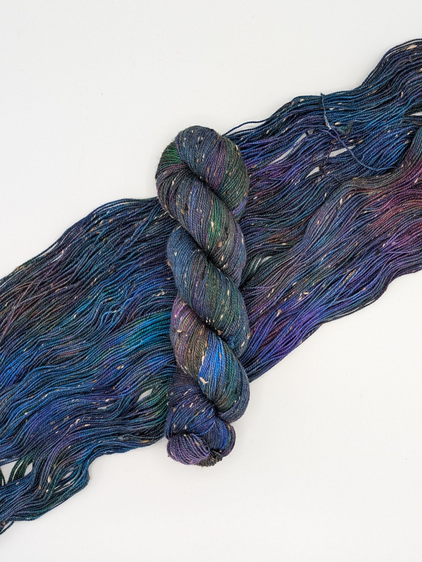 A skein of Galaxy Tweed Fingering weight yarn laying on a skein spread out to show it's jewel toned colors including blues, green, purples, greens.