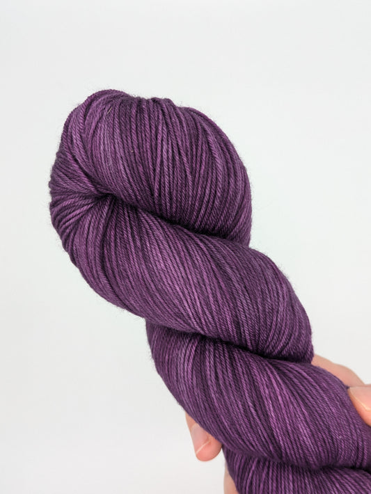 A hand holding a skein of Grape Jam Fingering weight yarn, showcasing a bright purple color