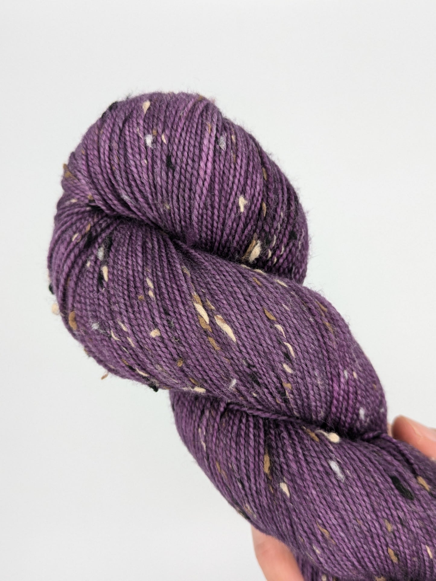 A hand holding a skein of Grape Jam Tweed Fingering weight yarn, showcasing a bright purple color