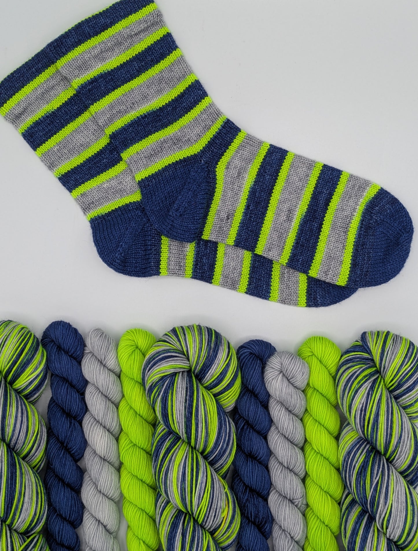 A pair of striped knit socks displayed above matching twisted yarn skeins on a clean white background. The socks and yarn feature navy blue, heather gray, and bright neon lime green stripes.