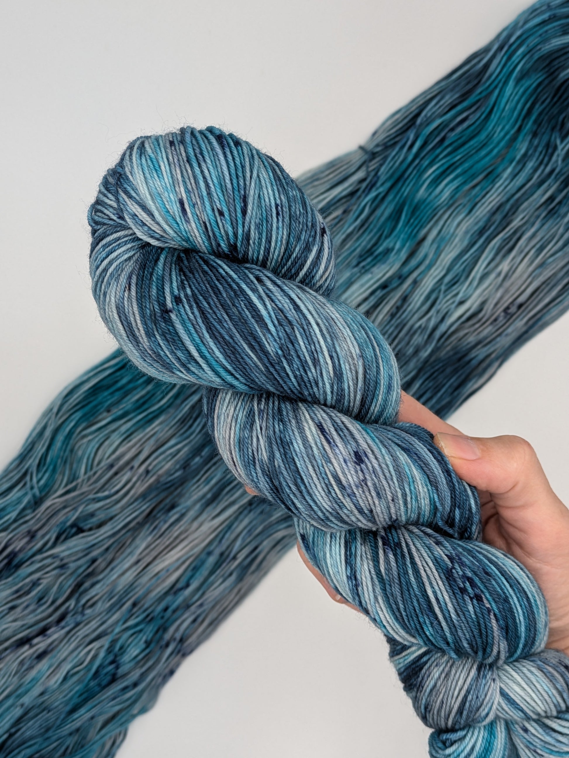 A hand holding a skein of MakerWool yarn in the color Winter's Chill, a variegated color with shades of blue and gray, topped with dark speckles.