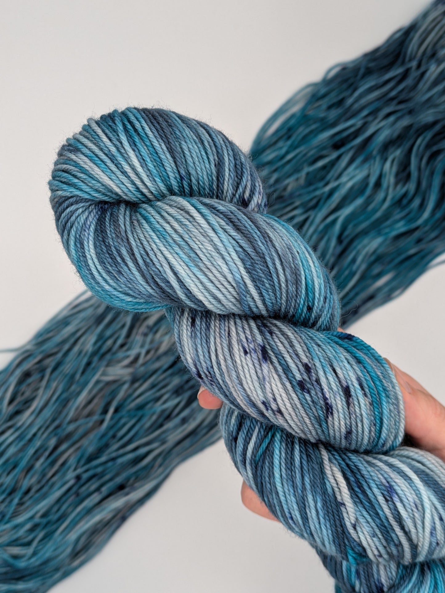 A hand holding a skein of MakerWool yarn in the color Winter's Chill, a variegated color with shades of blue and gray, topped with dark speckles.