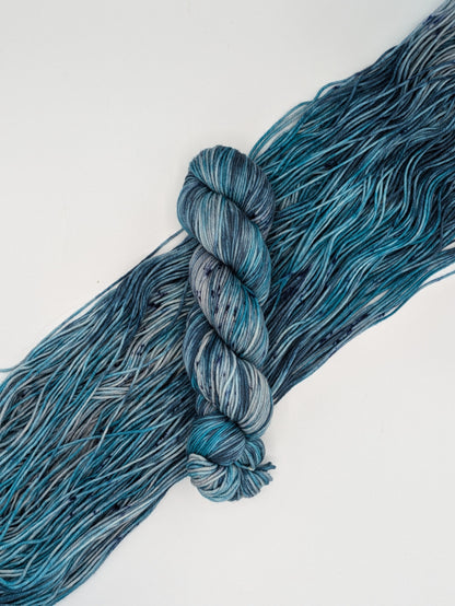 A skein of MakerWool yarn in the color Winter's Chill, a variegated color with shades of blue and gray, topped with dark speckles laying on top of a skein spread out to show the colors.