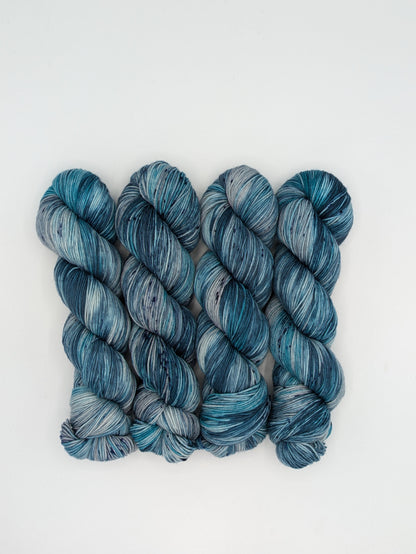 4 skeins of MakerWool Fingering Weight yarn in the color Winter's Chill, a variegated color with shades of blue and gray, topped with dark speckles.