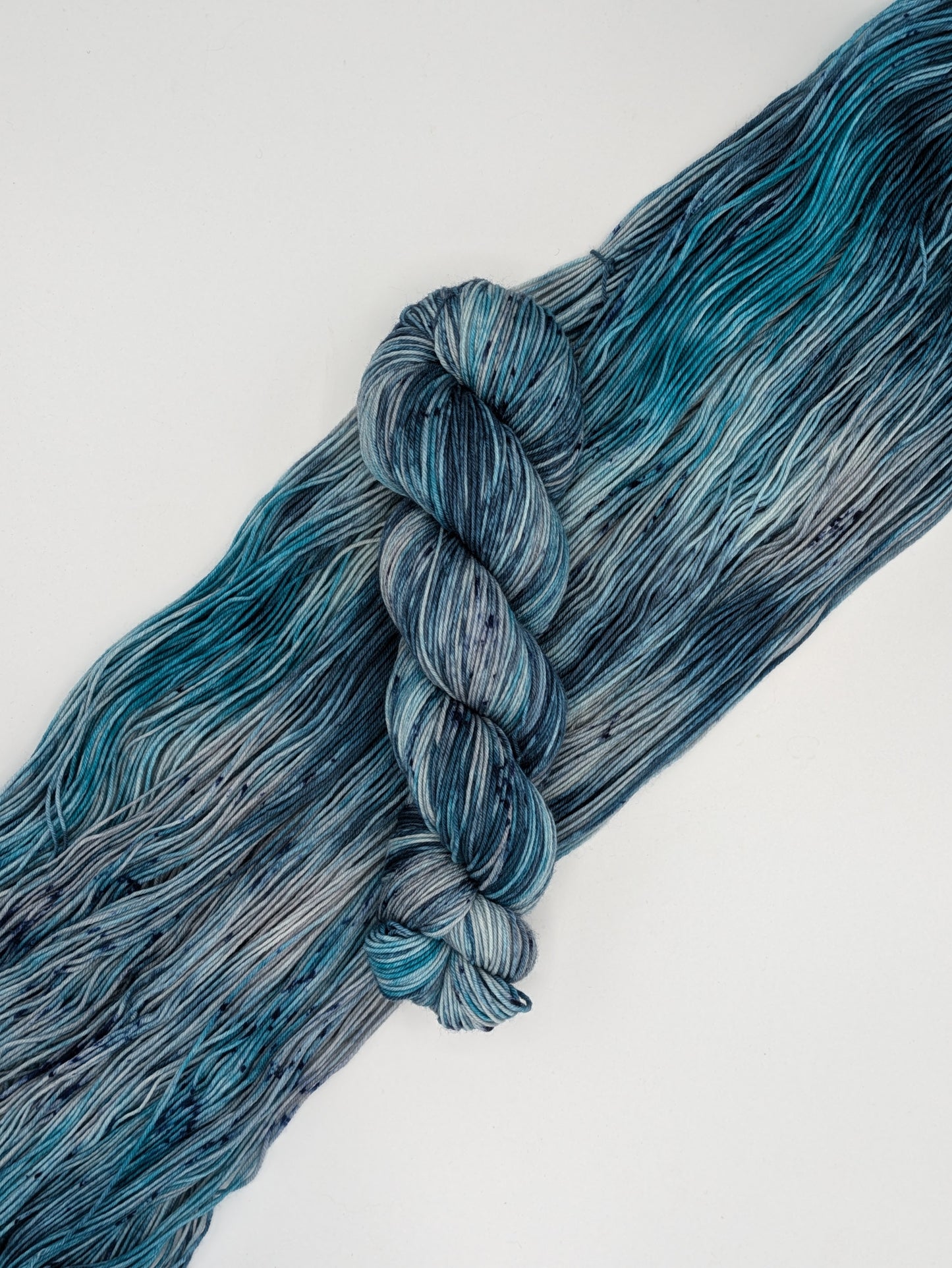 A skein of MakerWool yarn in the color Winter's Chill, a variegated color with shades of blue and gray, topped with dark speckles laying on top of a skein spread out to show the colors.