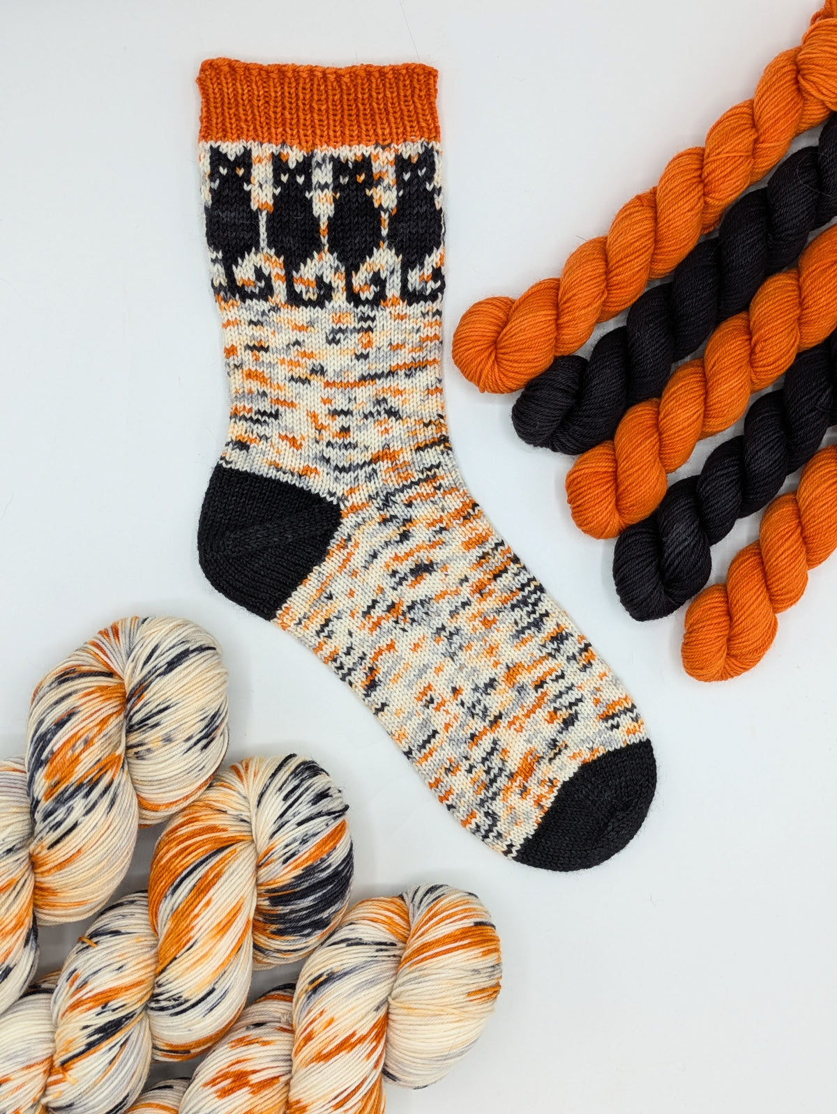 A single hand-knit sock with a black, orange, and cream variegated pattern is displayed on a white background, surrounded by matching variegated yarn skeins.
